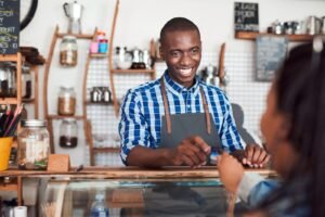 Smiling African male and small business owner, dressed in a checkered shirt and an apron, and listening to a customer talk about artificial intelligence